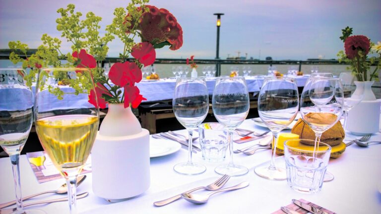 Lange gedekte tafel met een vaas met bloemen en wijnglazen vooraan en uitzicht op Amsterdam in de verte
